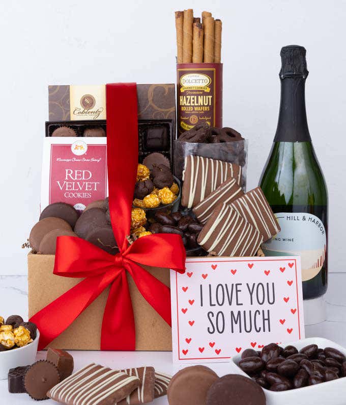 A gift box overflowing with treats including an assortment of chocolate-covered items like oreoes, almonds, graham crackers and pretzels along with red velvet cookies, hazelnut wafers, an 8 piece box of chocolates, a bottle of sparkling wine and an I Love