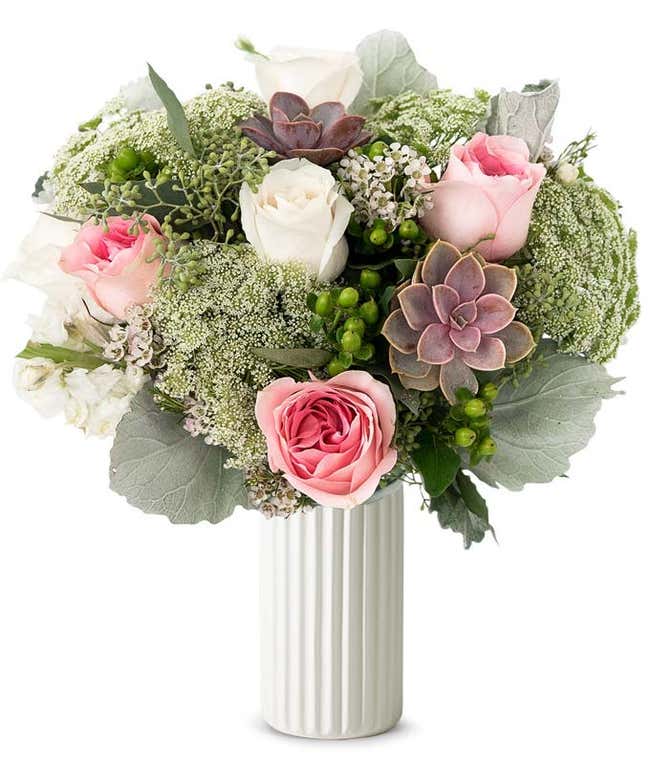 A floral arrangement featuring white roses, pink roses, succulents, white stock, hypericum berries, Queen Anne's lace, white waxflower, and seeded eucalyptus, with an optional glass vase.