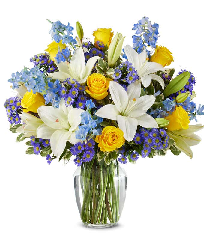 An elegant floral arrangement featuring white Asiatic lilies, yellow roses, blue delphinium, and purple Monte Casino, optionally arranged in a glass vase. This vibrant mix offers a stunning and colorful display.