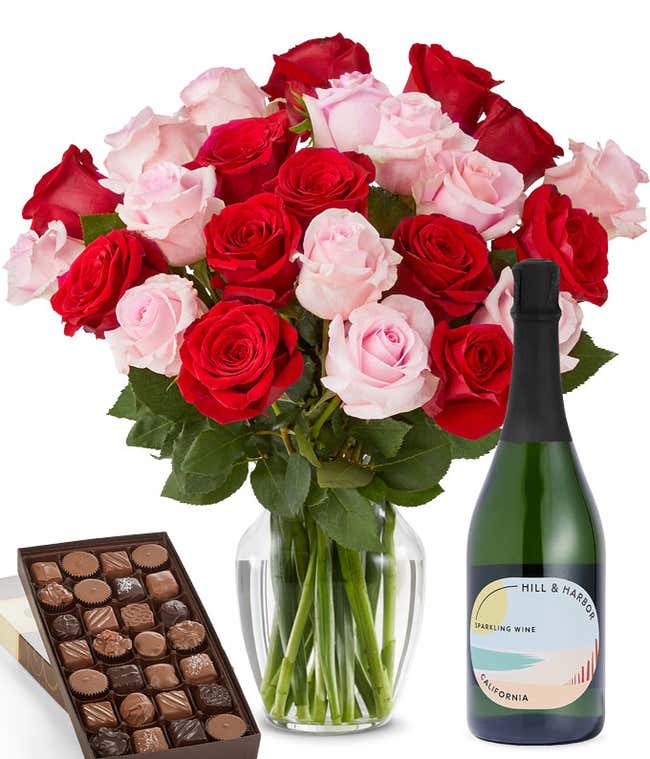 A bouquet of 24 red and pink roses arranged in a glass vase is displayed beside a bottle of Hill &amp; Harbor Sparkling Wine and a 1 pound box of assorted chocolates.