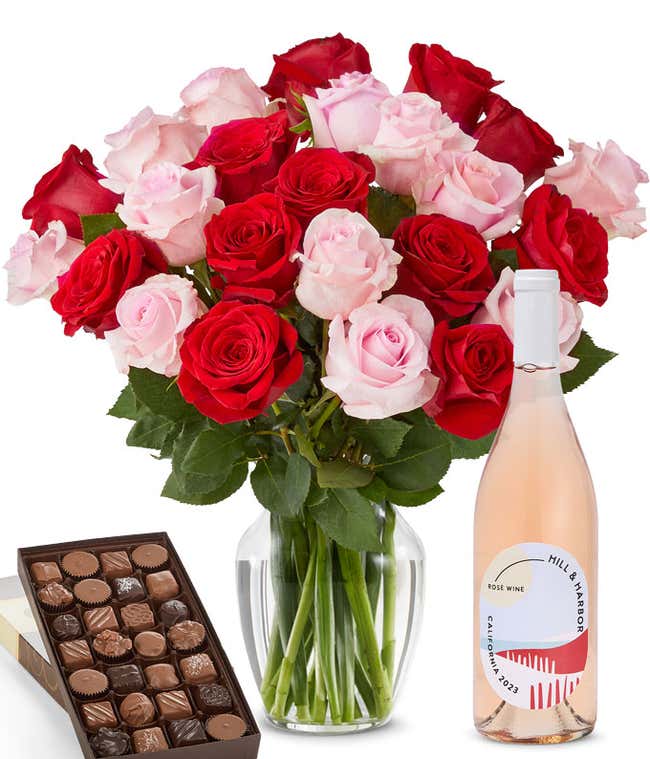 A bouquet of 24 red and pink roses arranged in a glass vase is displayed beside a bottle of Hill &amp; Harbor Ros&eacute; and a 1 pound box of assorted chocolates.