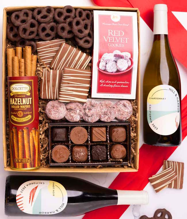 A gift box with chocolate pretzels, chocolate graham crackers, red velvet cookies, hazelnut wafer rolls, an 8 piece box of chocolates and 2 bottles of wine.