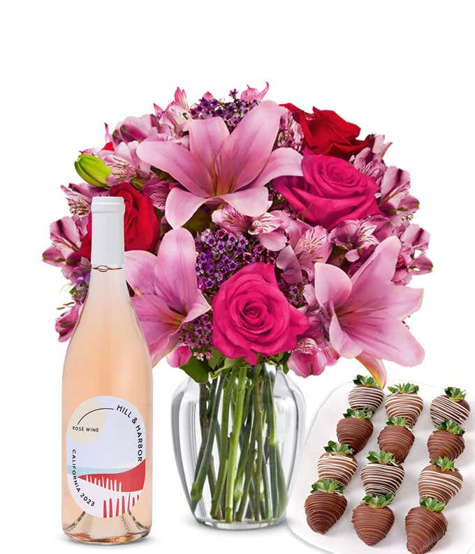 A mixed bouquet of pink and purple flowers in a clear glass vase is displayed with a bottle Hill & Harbor Rosé and 12 chocolate-covered strawberries.