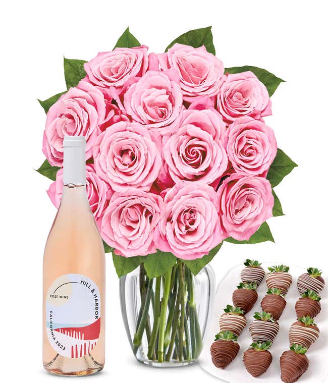 A bouquet of 1 dozen soft pink roses arranged in a clear glass vase is displayed beside a bottle of Hill &amp; Harbor Ros&eacute; and a neat assortment of 12 chocolate-covered strawberries.