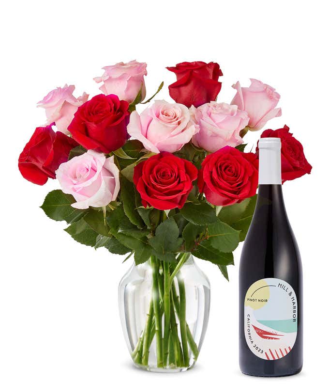 A mix bouquet of one dozen red and blush roses fills a clear glass vase and is paired with a bottle of Hill & Harbor California Pinot Noir.