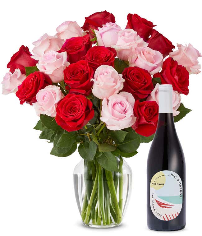 A mix bouquet of two dozen red and blush roses fills a clear glass vase and is paired with a bottle of Hill &amp; Harbor California Pinot Noir.
