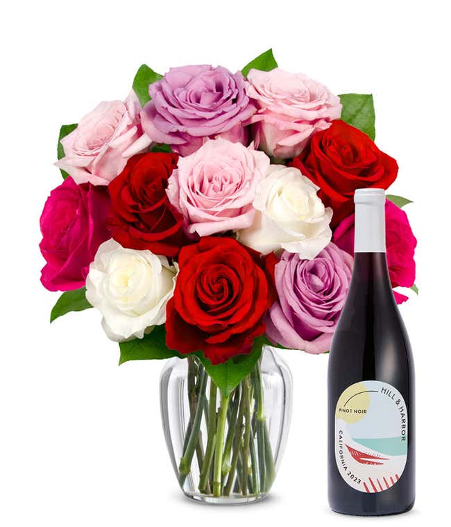 One dozen pink, purple and red roses arranged in a clear glass vase sits beside a bottle of Hill &amp; Harbor California Pinot Noir.