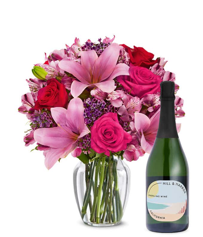 A bouquet of pink lilies, pink roses, and greenery arranged in a clear glass vase sitting next to a bottle of Hill & Harbor California Sparkling Wine.