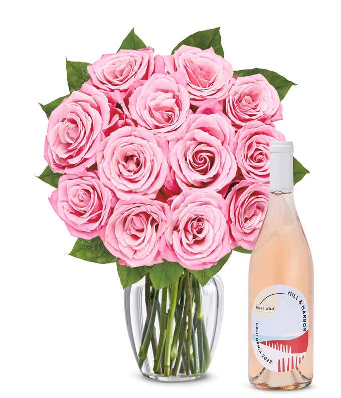 A bouquet of 12 soft pink roses arranged in a clear glass vase is displayed next to a bottle of Hill & Harbor California Rosé.