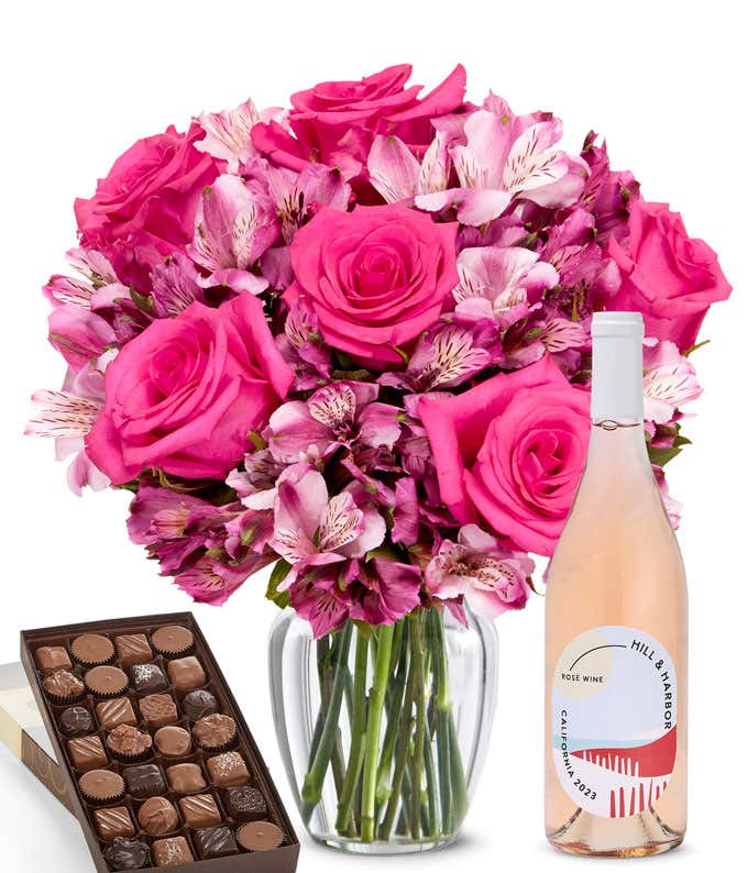 A bouquet of pink and roses and lilies in a clear glass vase is paired with a bottle of Hill & Harbor Rosé and a box of assorted chocolates.