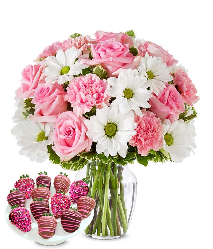 Deluxe bouquet of pink roses, pink carnations, and white daisy poms in a clear glass vase, paired with chocolate-covered strawberries decorated with pink drizzle and pink sprinkles.
