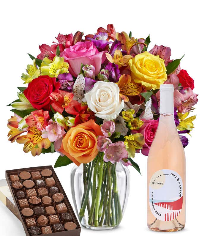 A colorful mixed bouquet of roses and alstroemeria flowers arranged in a clear vase is displayed beside a bottle of Hill & Harbor Rosé and a box of assorted chocolates.