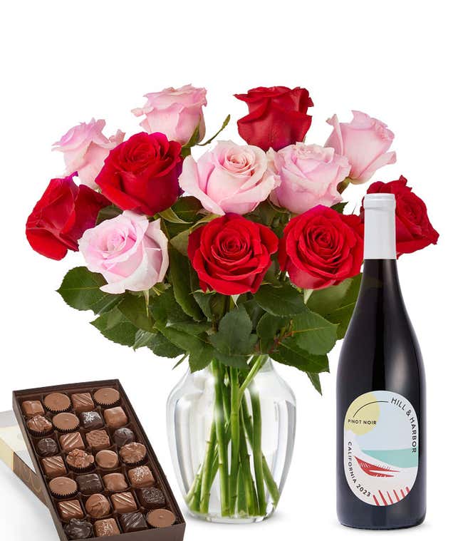 A bouquet of 12 red and pink roses arranged in a glass vase is displayed beside a bottle of Hill &amp; Harbor Pinot Noir and a 1 pound box of assorted chocolates.
