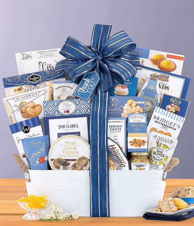 White gift basket with navy blue bow, filled with Ahmad English Tea, cookies, chocolates, hummus, cheese spread, pretzels, olives, and gourmet snacks, displayed with a small blue dish of treats.