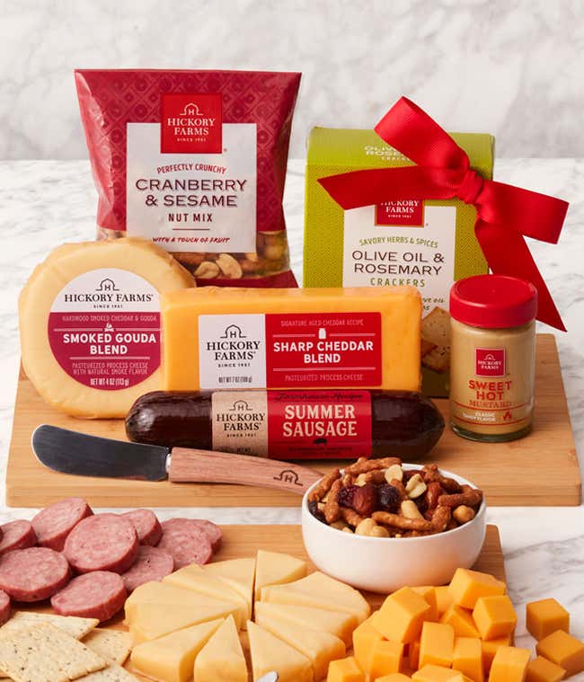 Gourmet gift set with Sharp Cheddar, Smoked Gouda, Olive Oil &amp; Rosemary Crackers, Cranberry &amp; Sesame Nut Mix, Farmhouse Summer Sausage, and Sweet Hot Mustard, displayed on a bamboo cutting board with a cheese spreader.