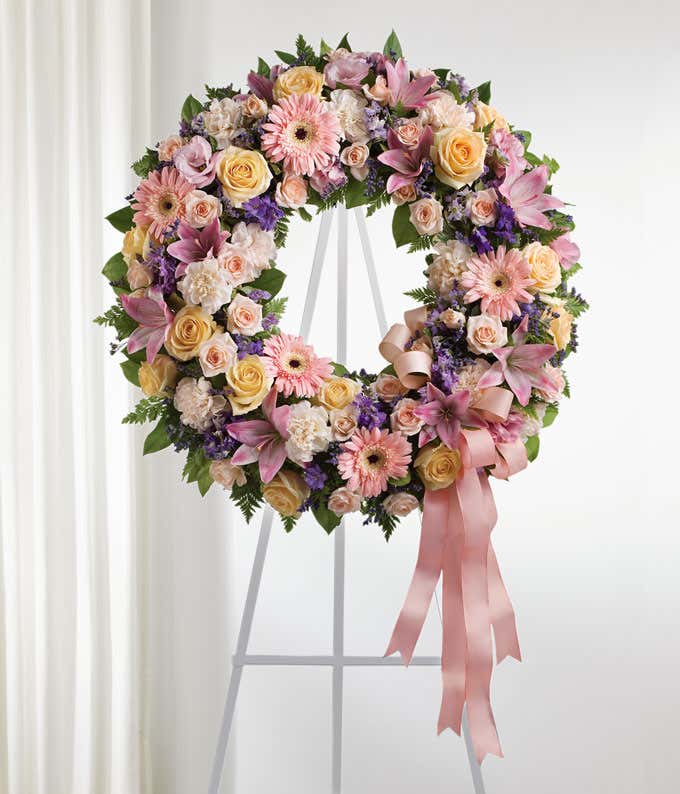 Funeral wreath with peach roses, pink lilies, peach carnations, green foliage, and a pink satin ribbon on a white metal easel.