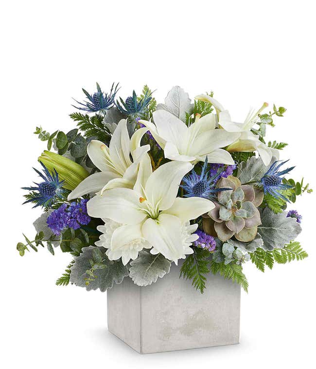 A floral arrangement in a square gray container featuring white lilies, purple thistle, green succulents, silver foliage, and seasonal greenery.
