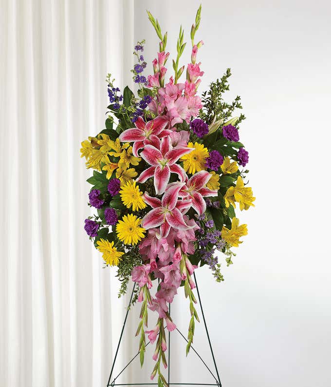 A standing funeral spray adorned with Pink Stargazer Lilies, Yellow Gerberas, Yellow Alstroemeria, Pink Gladioli, Purple Carnations, and Purple Larkspur, evoking a sense of grace, warmth, and reverence in remembrance of a beloved soul.