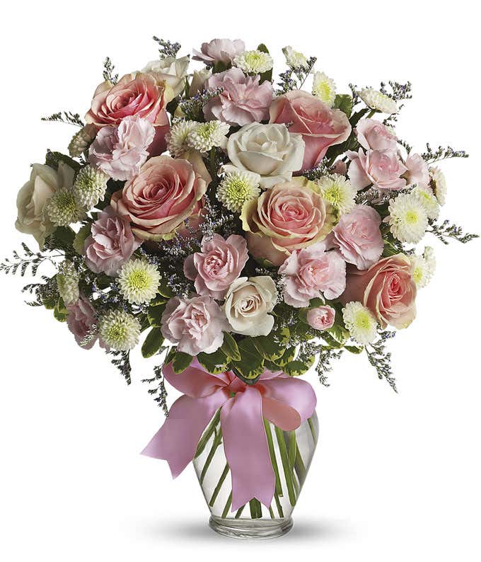 Floral arrangement featuring pink roses, white roses, mini carnations, and green poms in a clear glass vase with a bow.