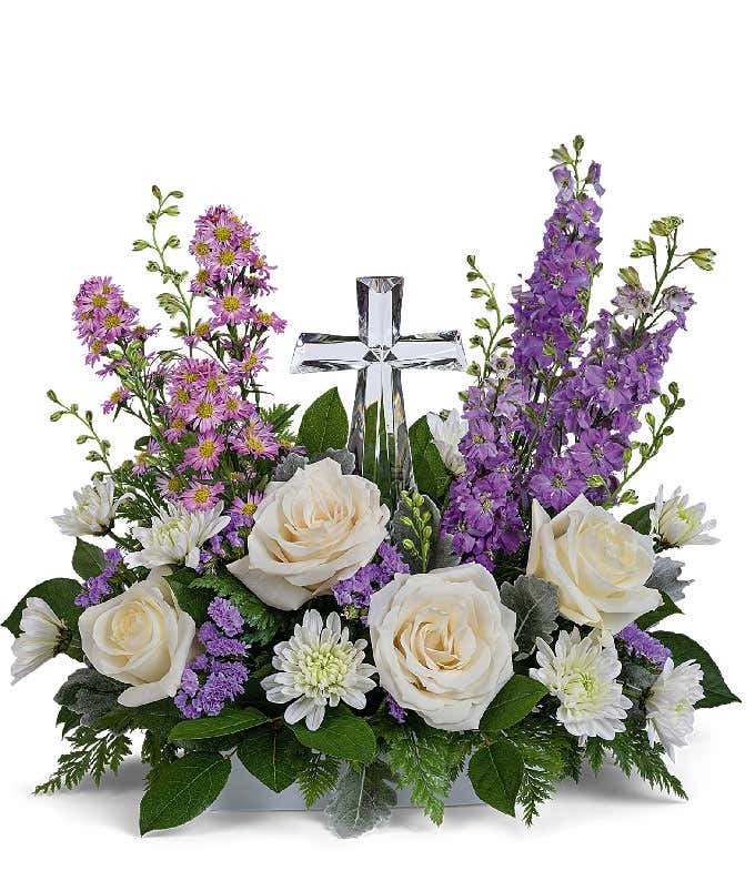A graceful floral arrangement showcasing crème roses, lavender larkspur, lavender asters, lavender statice, white mums, and floral greens, elegantly accented with a crystal cross. This bouquet blends soft, calming colors for a serene display.