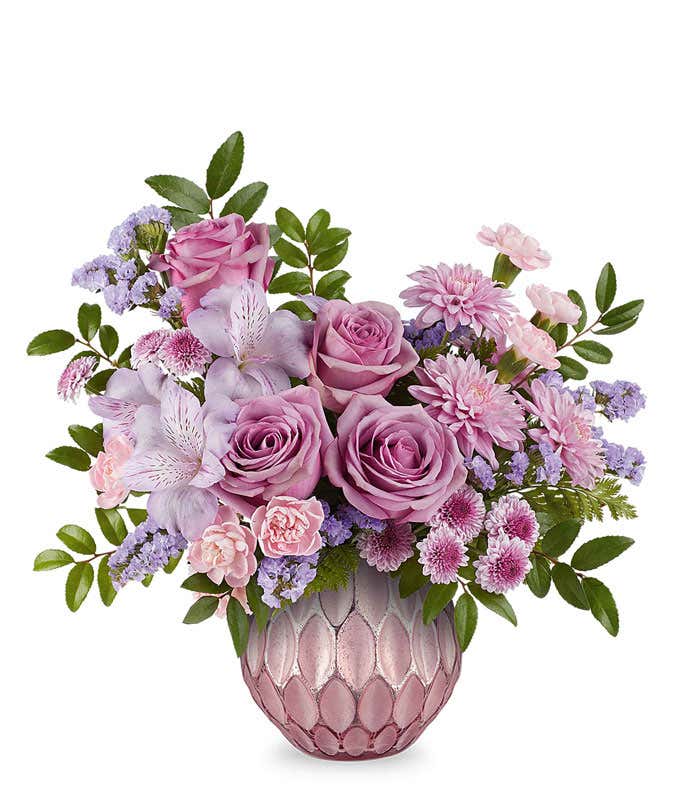 Lavender roses, lavender alstroemeria, miniature pink carnations, lavender button spray chrysanthemums, and lavender sinuata statice accented with greenery in a textured pink vase.