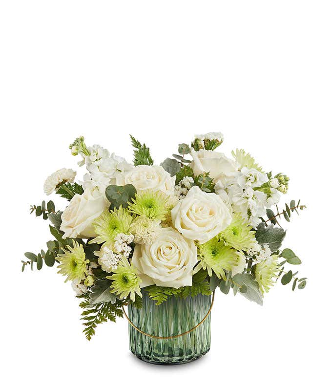 White roses, white stock, green button spray chrysanthemums, white cushion spray chrysanthemums, and assorted greenery arranged in a ribbed green glass vase