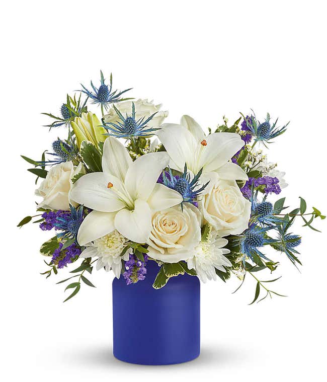 White lilies, white roses, white cushion spray chrysanthemums, blue eryngium, purple accent flowers, and greenery arranged in a matte cobalt blue cylinder vase.
