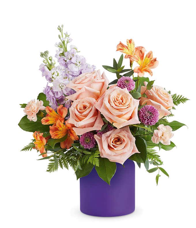 Floral arrangement with peach roses, orange alstroemeria, light lavender stock, lavender button poms, peach miniature carnations, and assorted greenery in a purple cylinder vase.