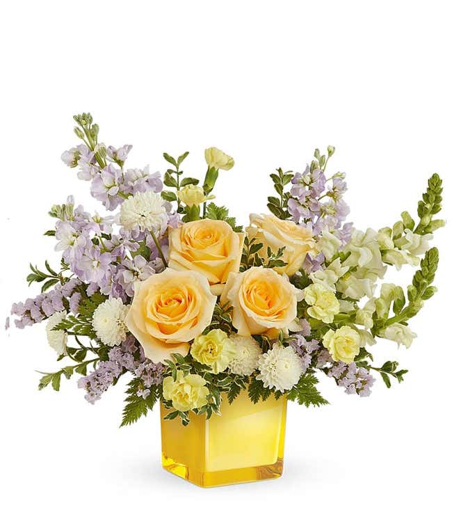 Yellow roses, mini yellow carnations, lavender stock, white button poms, white snapdragons, and floral greenery in a yellow cube vase 