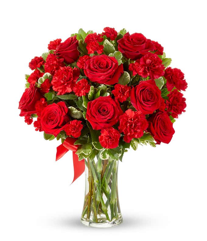 Floral arrangement featuring red roses, red carnations, red mini carnations, and a decorative red ribbon, florist delivered in a clear glass vase.