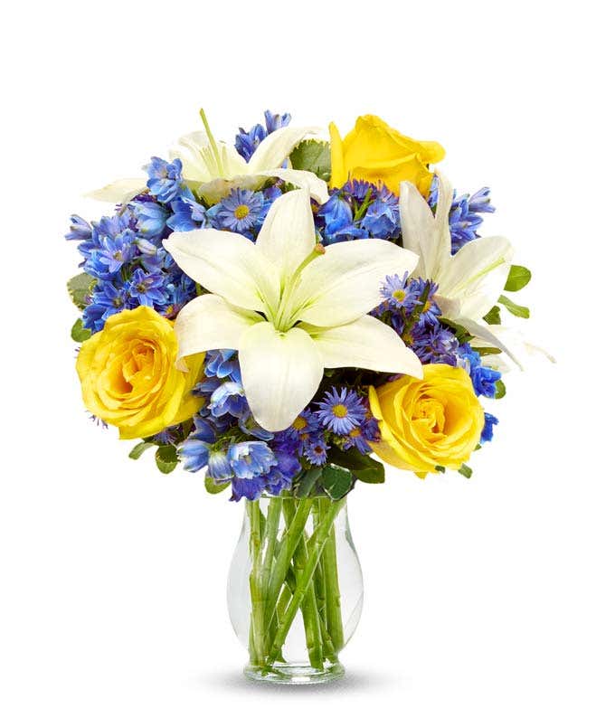Yellow roses, blue delphinium and white lilies in a clear glass vase