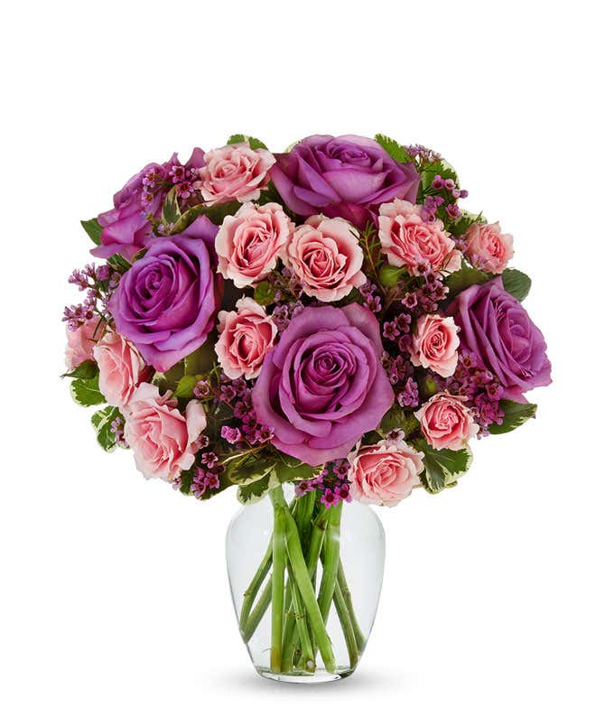 A vibrant bouquet of lavender roses, pink spray roses, and purple waxflower accented with lush greenery, arranged in a clear glass vase.