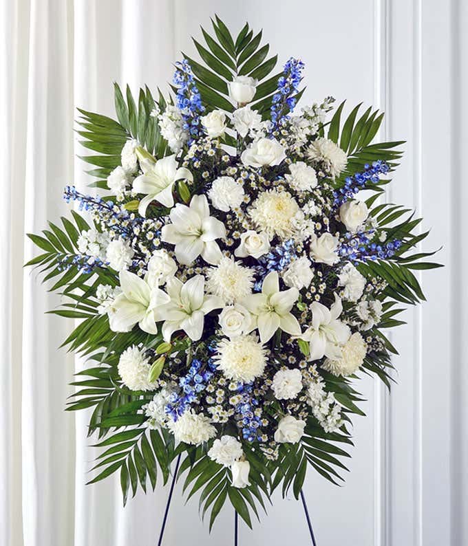 Standing spray arrangement featuring white lilies, roses, carnations, and poms, accented with blue delphinium, white monte casino, and palm fronds on a display easel. 