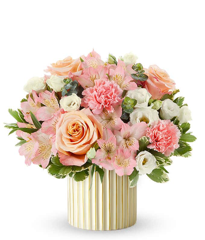 Arrangement of peach roses, pink alstroemeria, pink carnations, white blooms, and variegated greenery in a ribbed ivory planter with gold accents.