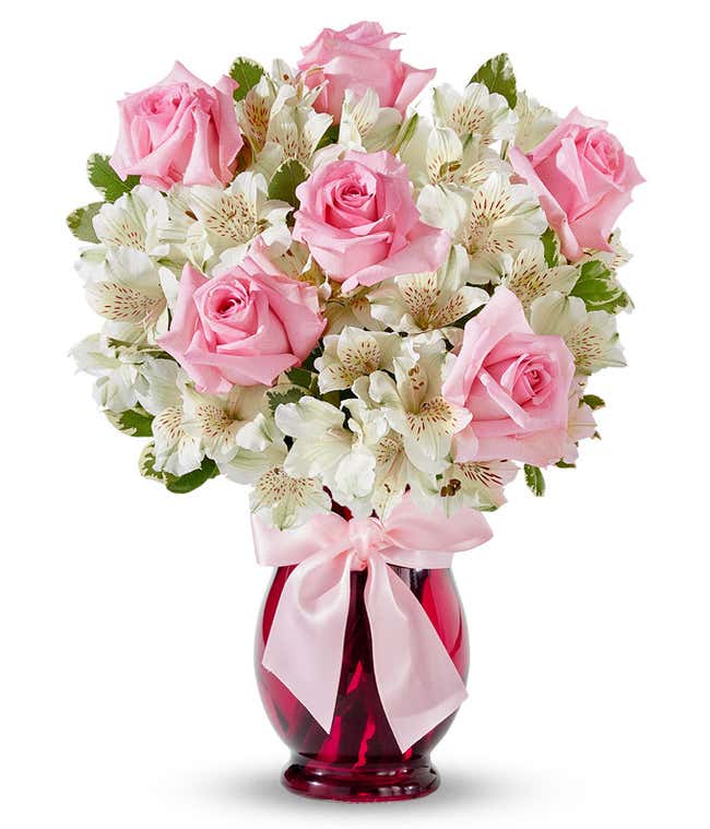 Bouquet of light pink roses and white alstroemeria with red speckled centers arranged in a deep pink glass vase tied with a pale pink satin bow.