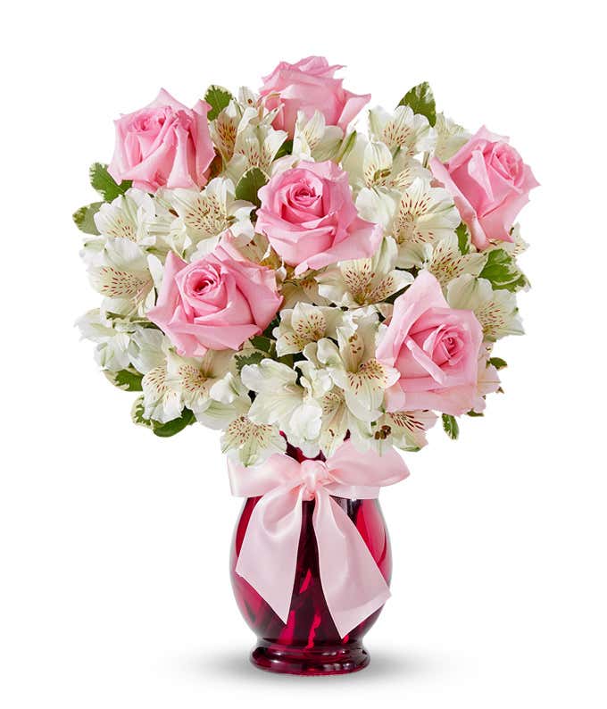 Bouquet of light pink roses and white alstroemeria with red speckled centers arranged in a deep pink glass vase tied with a pale pink satin bow.
