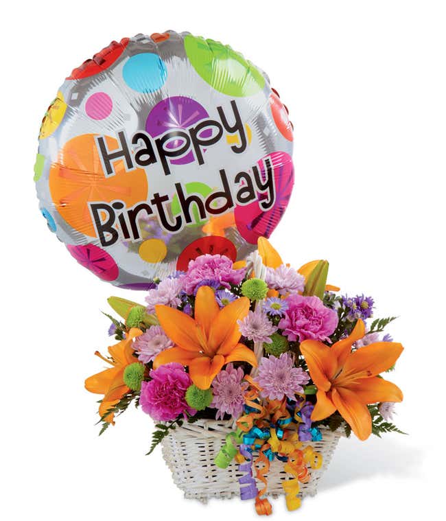 Festive birthday flower arrangement with orange lilies, pink carnations, purple poms, green button poms, and a colorful &ldquo;Happy Birthday&rdquo; balloon in a white basket.