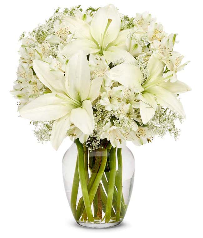 Clear glass vase filled with white lilies, white alstroemeria, and delicate white filler flowers, arranged in a full, rounded bouquet with visible green stems.