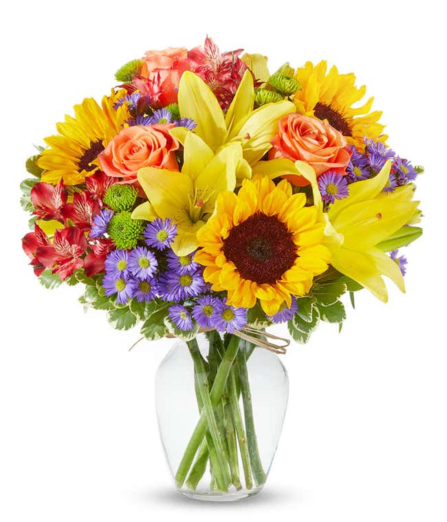 An arrangement of sunflowers, yellow lilies, orange roses, alstroemeria, Monte Casino, and green poms, in a clear glass vase.