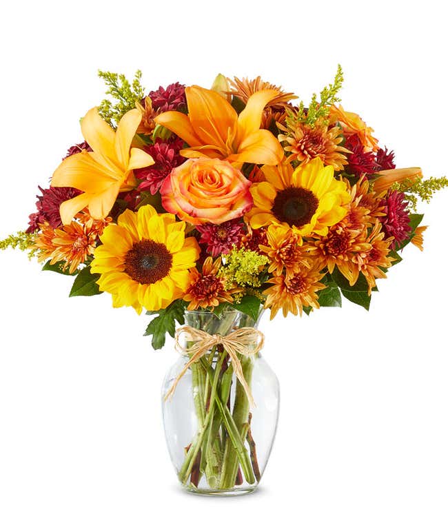 Bright autumn bouquet with sunflowers, orange lilies, orange roses, burgundy poms, yellow solidago, and butterscotch daisies in a clear glass vase tied with a raffia bow.