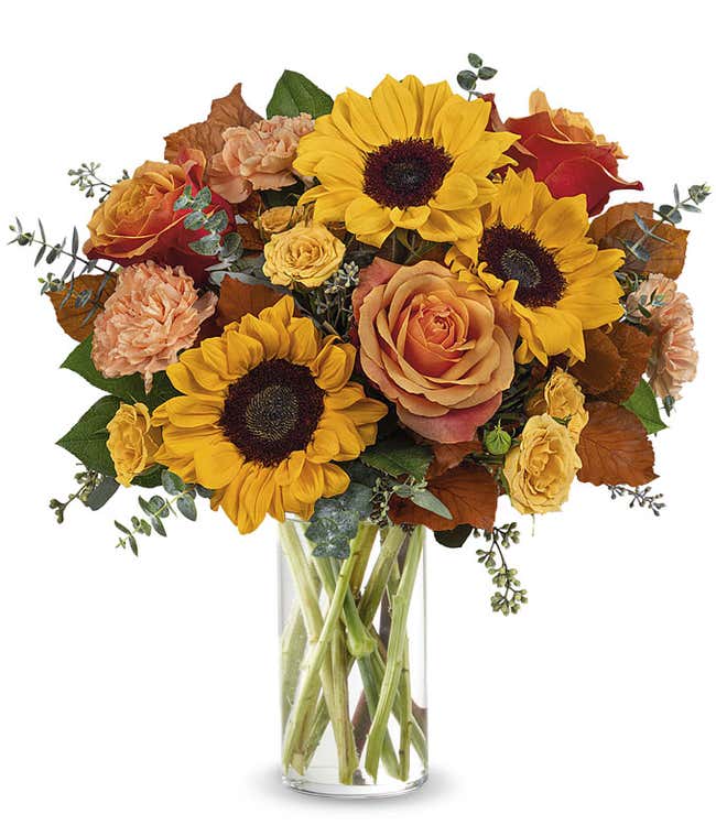 Fall bouquet with sunflowers, orange roses, peach carnations, yellow spray roses, seeded eucalyptus, and autumn oak leaves in a tall clear glass vase.