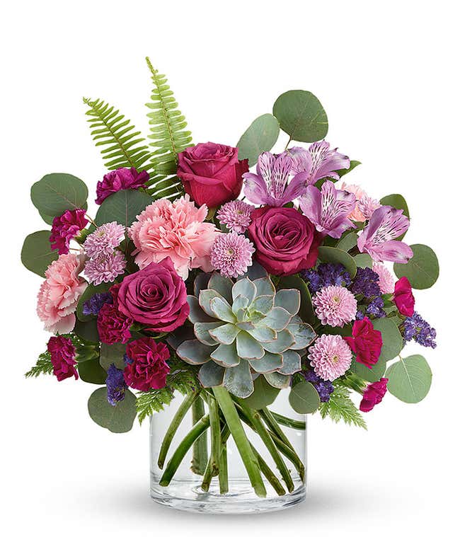Floral arrangement featuring a succulent plant, pink carnations, purple mini carnations, dark pink roses (in deluxe and premium versions), purple statice, purple alstroemeria, lavender button mums, seasonal greenery in a glass vase