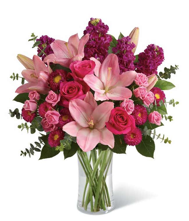 Pink lilies, hot pink roses, pink spray roses, magenta Matsumoto asters, and purple stock arranged with eucalyptus and greenery in a clear glass vase, creating a bold and elegant pink floral bouquet.