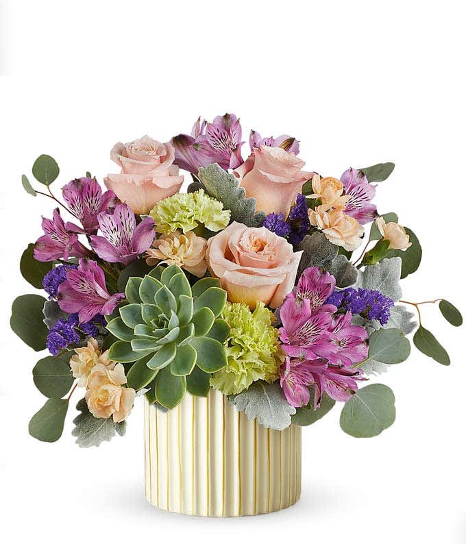 Floral arrangement with blush roses, purple alstroemeria, green carnations, small peach blooms, a large green succulent, and eucalyptus in a ribbed ivory planter with gold accents.