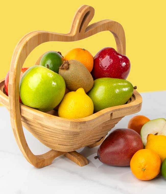 A bamboo fruit shaped like an apple containing pears, oranges, kiwis, limes, lemmons and apples with some cut fruit on the counter next to the bowl.