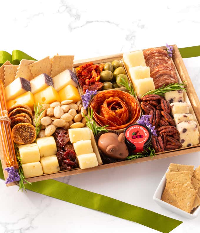 Charcuterie board with assorted cheeses, salami slices, sun-dried tomatoes, green olives, fig and almond clusters, dried cherries, pecans, flatbread crackers, blueberry Stilton, sour cherry spread, a chocolate mouse, and floral accents.