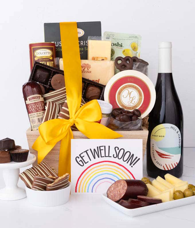 A wooden basket filled with chocolates, cheese, sausage, crackers, crisps, and snacks, paired with a bottle of red wine and a rainbow Get Well Soon card, with chocolates and charcuterie displayed.