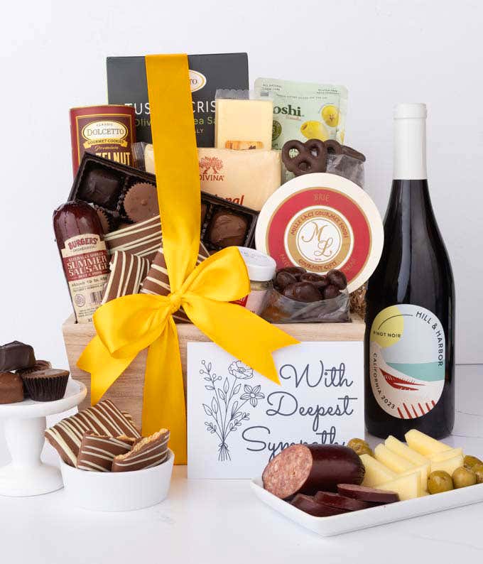 A wooden basket filled with chocolates, cheese, sausage, crackers, crisps, and snacks, paired with a bottle of red wine and a With Sympathy card, with chocolates and charcuterie displayed.