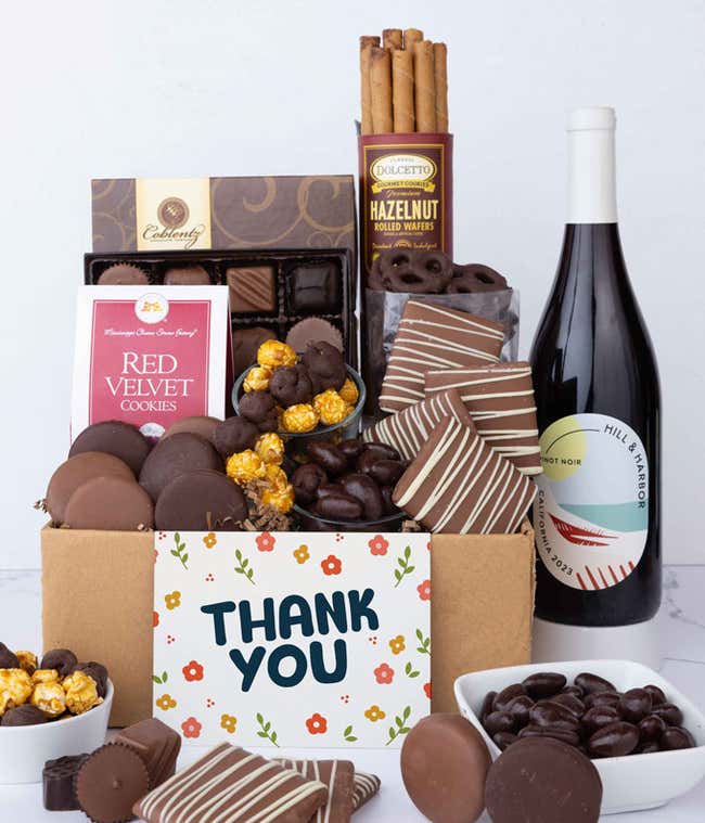 A gift box overflowing with treats including an assortment of chocolate-covered items like oreoes, almonds, graham crackers and pretzels along with red velvet cookies, hazelnut wafers, an 8 piece box of chocolates and a bottle of red wine.