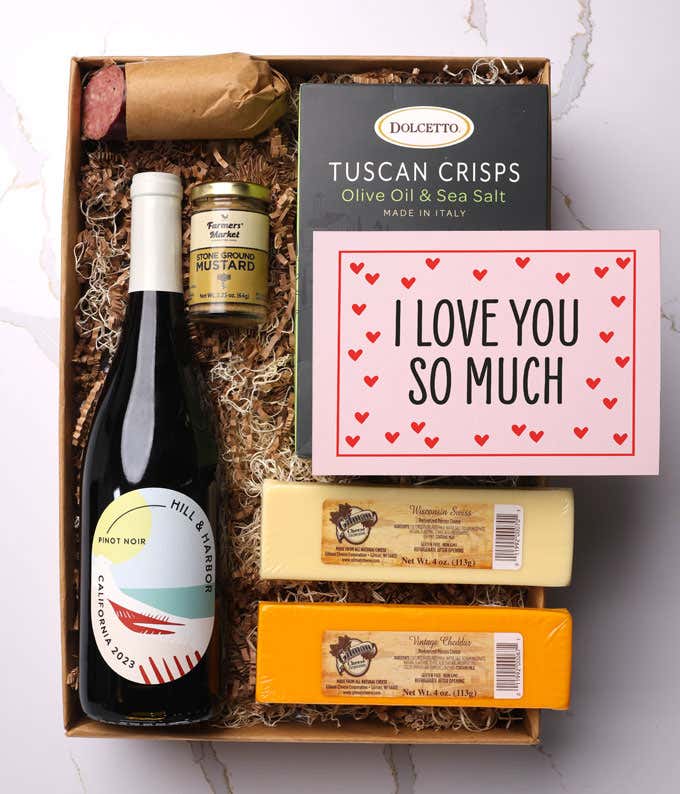 A beautifully arranged gift box featuring a bottle of Hill & Harbor Pinot Noir, two hard cheeses, a jar of stone-ground mustard, gourmet crackers, savory summer sausage, and a pink card with hearts that reads 'I Love You So Much'